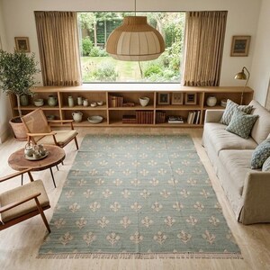 May include: A rectangular area rug featuring a muted sage green background with a repeating cream-colored floral motif and short fringe along the edges. The rug is positioned in the center of a living room with light wood flooring and furniture.