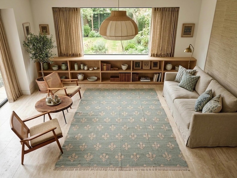 May include: A rectangular area rug featuring a muted sage green background with a repeating cream-colored floral motif and short fringe along the edges. The rug is positioned in the center of a living room with light wood flooring and furniture.