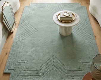 May include: A rectangular sage green area rug featuring a raised geometric pattern of concentric chevron lines. The rug has a textured, tufted surface and is positioned on a light wood floor, centered beneath a round white marble coffee table.