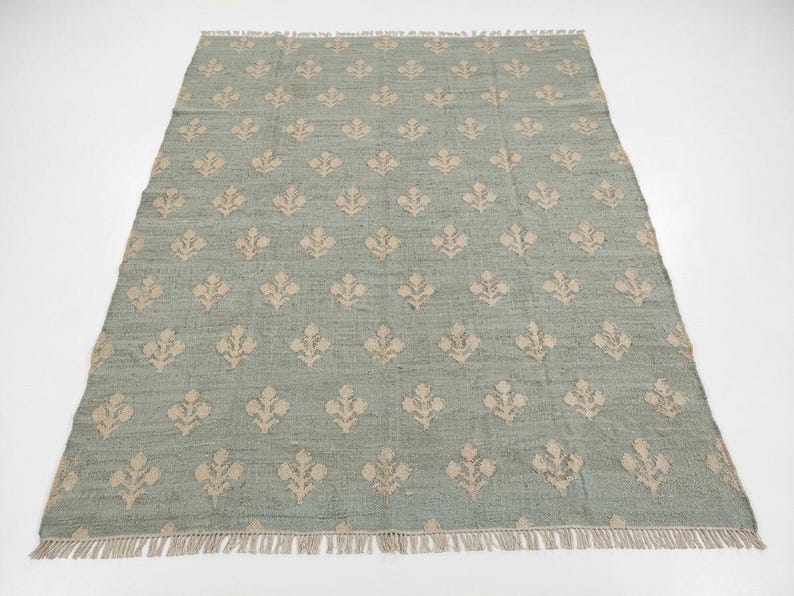 May include: A rectangular area rug featuring a muted sage green base color with a repeating pattern of small, light beige floral motifs arranged in a grid. The rug has a textured, woven appearance and includes short fringe along both ends.