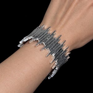 Puede incluir: Una pulsera brazalete ancha de tono plateado con una textura dentada similar a púas que se extiende por los bordes superior e inferior. La superficie metálica tiene un acabado reflectante con surcos profundos y puntas afiladas e irregulares.