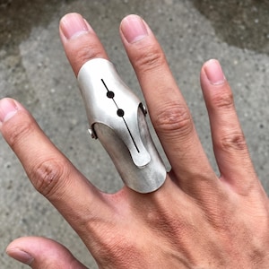 May include: A silver-toned metal finger splint ring featuring a smooth, curved surface with a central vertical slit and two small circular cutouts. The piece is constructed with a hinged design, secured by small rivets on the sides, covering the finger.