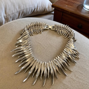 May include: A silver-toned statement necklace featuring a series of curved, elongated, spike-like metal segments arranged in a circular collar formation. The piece includes a metallic box clasp closure at the back to secure the necklace around the neck.