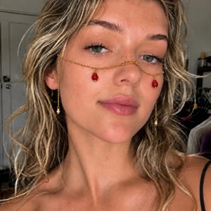 May include: A gold-toned chain face accessory featuring a delicate link design that rests across the bridge of the nose. Two vibrant red teardrop-shaped charms hang from the chain, while smaller white beads dangle from the ends near the ears.