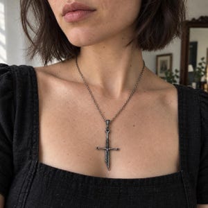 以下が含まれることがあります： クロスガード付きの剣の形をしたペンダントが付いたシルバー調の金属製ネックレス。ペンダントは繊細な丸いリンクチェーンから吊り下げられています。剣のデザインには質感のあるハンドルと尖った刃が含まれており、暗い酸化仕上げのパティナが施され、金属細工の複雑なディテールを際立たせています。