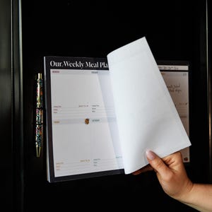 May include: A magnetic weekly meal planner notepad with a black border and white pages featuring the text Our Weekly Meal Plan. The planner includes sections for days of the week, cooking time, recipes, and notes, accompanied by a floral patterned pen.