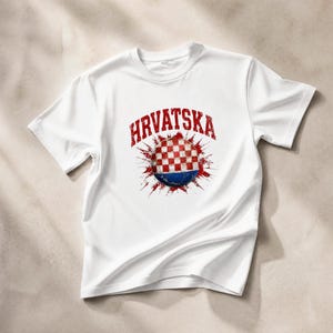 May include: White t-shirt featuring the word "HRVATSKA" in red above a graphic of a Croatian flag design. The graphic is a red and white checkered ball with a blue base, surrounded by red splatter effects.