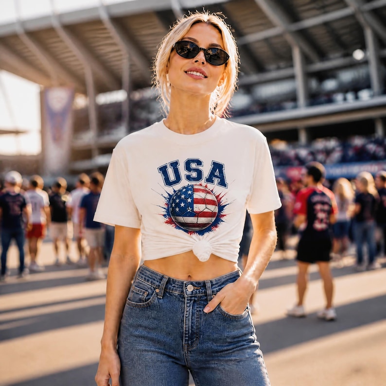 May include: Cream-colored t-shirt with the word "USA" in blue lettering above a soccer ball design featuring the American flag. The shirt is knotted at the waist and paired with blue jeans. The image is taken outdoors.