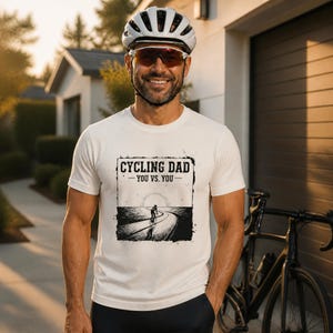 Könnte beinhalten: Ein weißes kurzärmeliges T-Shirt mit einem schwarzen Grafikdruck eines Radfahrers auf einer Straße und dem Text "CYCLING DAD - YOU VS. YOU -", zentriert in einem quadratischen Rahmen. Das Design zeigt eine untergehende Sonne und abgenutzte Ränder.