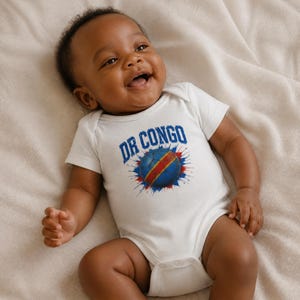 Könnte beinhalten: Ein weißer kurzärmeliger Babybody mit einem Grafikdruck eines Fußballs in den Farben der Flagge der Demokratischen Republik Kongo, umgeben von blauen und roten Farbspritzern, mit dem Text DR CONGO in blauen Blockbuchstaben.