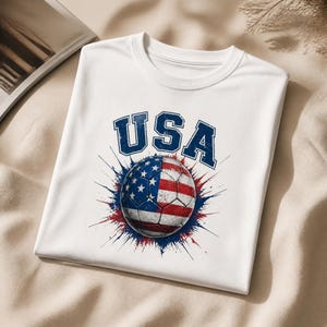 May include: White t-shirt featuring a soccer ball design with the American flag and the word "USA" in blue. The soccer ball is red, white, and blue, with a star pattern. The design has a distressed, vintage look.