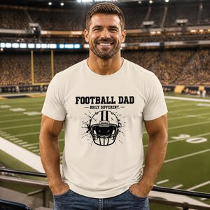 Könnte beinhalten: Ein cremefarbenes Kurzarm-T-Shirt mit einem schwarzen Grafikdruck eines Football-Helms und Farbspritzern. Der Text FOOTBALL DAD und BUILT DIFFERENT ist auf der Vorderseite des T-Shirts in schwarzer Farbe aufgedruckt.