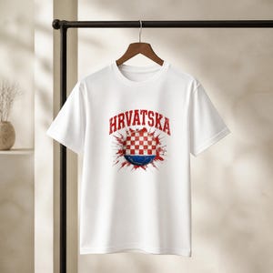 May include: White t-shirt with the word "HRVATSKA" in red, arched above a graphic. The graphic features a red and white checkered pattern, with a blue and red splash design. The t-shirt is hanging on a wooden hanger.