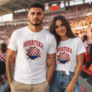 May include: Two people wearing white t-shirts with the word "HRVATSKA" in red, above a Croatian flag design. The shirts are in a stadium setting, suggesting a sports or national pride theme.