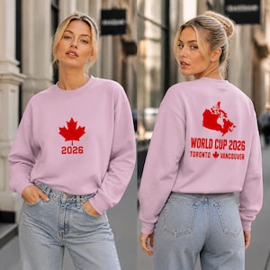 Könnte beinhalten: Ein hellrosa Sweatshirt mit einem roten Ahornblatt und der Zahl 2026 auf der Vorderseite. Auf der Rückseite des Sweatshirts befindet sich eine rote Karte von Kanada mit dem Text "WORLD CUP 2026" und den Städten "TORONTO" und "VANCOUVER".