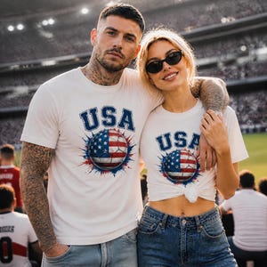 May include: Two people wearing white t-shirts with the word "USA" in blue block letters above a soccer ball design featuring the American flag. The soccer ball is red, white, and blue with a starburst effect. They are in a stadium.