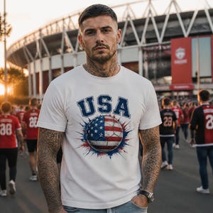 May include: White t-shirt with "USA" in blue lettering above a soccer ball design featuring the American flag. The shirt is worn by a person in a crowd, likely at a sporting event. The background includes a stadium and other people.