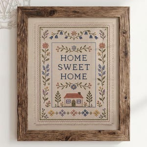 Puede incluir: Un bordado en punto de cruz enmarcado sobre tela beige con el texto HOME SWEET HOME en letras azules. El diseño incluye una pequeña casa, bordes florales en azul, rojo y verde, y patrones geométricos decorativos en un marco de madera rústica.