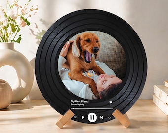 Op de afbeelding: Een zwarte, ronde fotolijst die lijkt op een vinylplaat. De lijst bevat een foto van een persoon en een hond. De tekst "My Best Friend Forever My Baby" wordt weergegeven, samen met mediabesturingspictogrammen. De lijst wordt ondersteund door twee houten standaards.