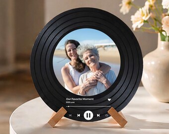 Op de afbeelding: Zwarte, ronde fotolijst in de vorm van een vinylplaat. Toont een foto van twee vrouwen. De tekst "Our Favorite Moment With Love" en bedieningselementen voor de muziekspeler zijn zichtbaar. De lijst wordt ondersteund door een houten standaard.
