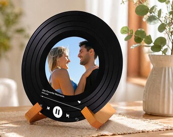 Op de afbeelding: Zwarte fotolijst in de vorm van een vinylplaat met een foto van een stel. De lijst heeft een houten standaard en heeft een muziekspelerinterface met de tekst "Our Favorite Moment With Love".