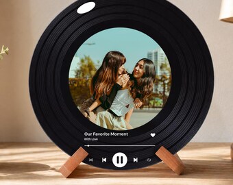 Op de afbeelding: Zwarte fotolijst in de vorm van een vinylplaat met een foto van twee personen. De lijst heeft een houten standaard en toont de tekst "Our Favorite Moment" en een muziekspelerinterface.
