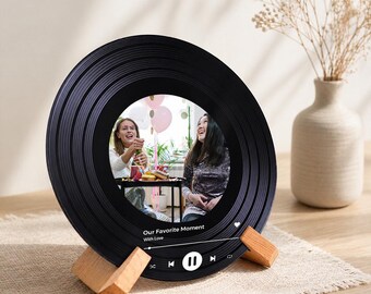 Op de afbeelding: Een zwarte fotolijst in de vorm van een vinylplaat met een foto van twee vrouwen. Het frame wordt ondersteund door twee kleine houten standaards. De tekst "Our Favorite Moment With Love" staat onderaan het frame.