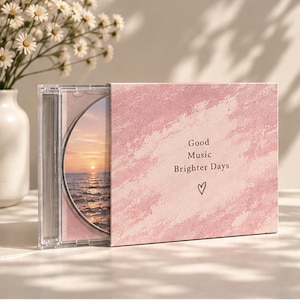 Może przedstawiać: Przezroczyste plastikowe etui na płytę CD z różową, teksturowaną obwolutą z napisem Good Music Brighter Days oraz małą ikoną serca. Widoczna wkładka płyty przedstawia zachód słońca nad oceanem ze spokojnym morzem i jasnym horyzontem.