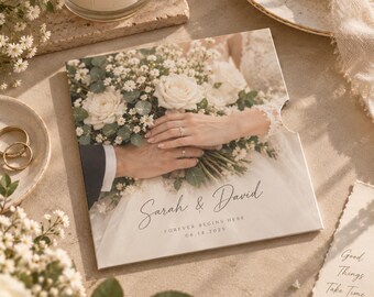 Op de afbeelding: Een vierkante trouwkaart met een foto van twee handen rustend op een wit bloemenboeket. De tekst luidt Sarah & David, Forever Begins Here, en 14.06.2025. De kaart is voorzien van een halfronde uitsparing aan de rechterkant.