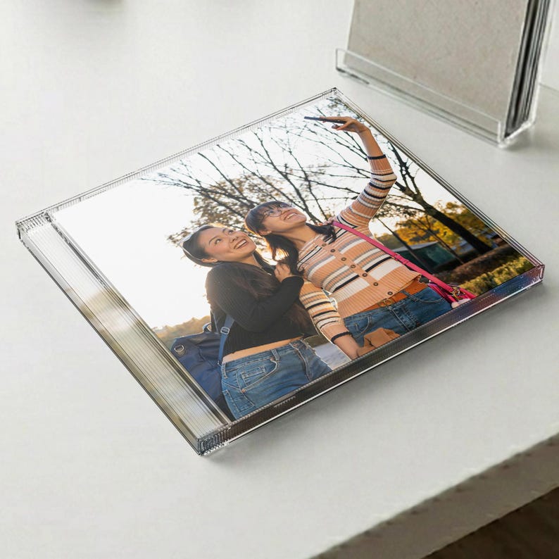 May include: A square clear acrylic photo block displaying a printed image of two individuals outdoors. One individual wears a black long-sleeved top and blue jeans, while the other wears a striped long-sleeved shirt and blue jeans, holding a phone.
