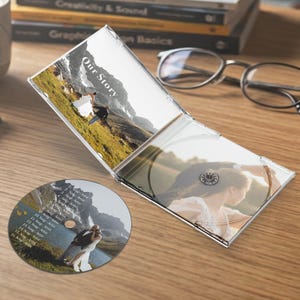 Puede incluir: Una caja de CD transparente con un CD dentro y un CD separado sobre una superficie de madera. La caja tiene una foto de una pareja en una montaña, con el texto "Our Story". El CD separado tiene una foto de una pareja junto a un lago.