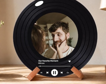 Op de afbeelding: Zwarte fotolijst in de vorm van een vinylplaat met een foto van een stel. De lijst heeft een houten standaard en bevat de tekst "Our Favorite Moment With Love" met bedieningselementen voor de muziekspeler.