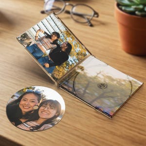 May include: A clear plastic CD jewel case containing a printed photograph of two people standing outdoors, accompanied by a circular disc featuring a close-up portrait of the same two individuals, both displayed on a wooden surface.