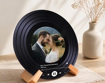 Puede incluir: Un marco de fotos negro con forma de disco de vinilo con una foto de boda en su interior. El marco está sostenido por dos soportes de madera. La foto muestra a una pareja abrazándose. El texto "Our Favorite Moment With Love" se muestra en la parte inferior.