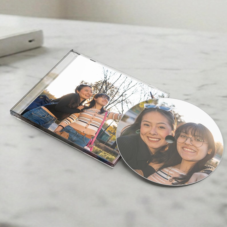 May include: A clear plastic CD jewel case and a circular CD disc featuring a printed photograph of two individuals smiling outdoors. One individual wears a black long-sleeve top and blue jeans, while the other wears a striped long-sleeve shirt.