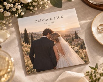 Op de afbeelding: Een vierkante trouwkaart met een foto van een koppel dat bij zonsondergang in een veld staat. De tekst OLIVIA & JACK en 09.14.2024 is in het zwart bovenaan gedrukt. De kaart heeft een gladde afwerking en een kleine halfronde duiminkeping.