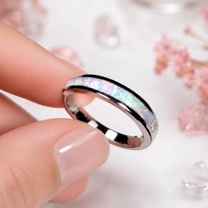 Puede incluir: Un anillo de metal plateado pulido con una incrustación central de ópalo iridiscente. La incrustación muestra un espectro de colores que reflejan la luz, incluyendo rosa, azul y verde, engastado en una banda lisa y reflectante.