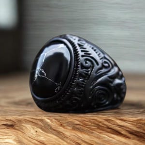 May include: A black ring featuring a large, oval-shaped black stone with white veining set into a dark metal band. The band is decorated with intricate, raised scrollwork and carved patterns that extend around the sides of the ring.