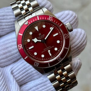 Peut inclure: Une montre Tudor Black Bay en acier inoxydable avec un cadran rouge et une lunette tournante rouge assortie. La montre présente un bracelet en métal à cinq maillons, des index blancs luminescents et le texte Tudor Geneve et Master Chronometer.