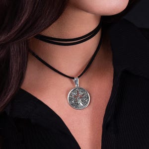 May include: A silver pendant necklace featuring a detailed tree of life design. The pendant hangs from a black cord necklace. The model is wearing a black collared shirt.