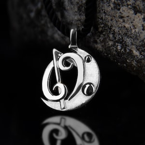 May include: Silver-toned pendant featuring a musical treble clef and a bass clef design. The pendant is circular with three small, round accents and hangs from a black cord. The background is dark with a stone texture.