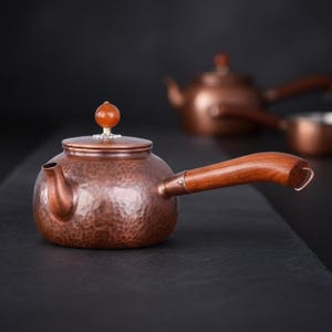 Könnte beinhalten: Eine Teekanne aus gehämmertem Kupfer mit einem seitlichen Holzgriff und einem Deckel, der mit einem kleinen, runden, bernsteinfarbenen Stein verziert ist. Die Kanne hat einen kurzen, gebogenen Ausguss und eine texturierte, gehämmert wirkende Oberfläche.