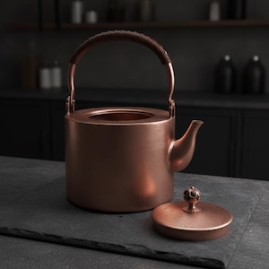 Puede incluir: Una tetera cilíndrica de cobre con un acabado metálico liso, que presenta un pico curvo y un asa alta y arqueada envuelta en material marrón oscuro. Una tapa circular a juego con un pomo decorativo se encuentra junto al cuerpo de la tetera.