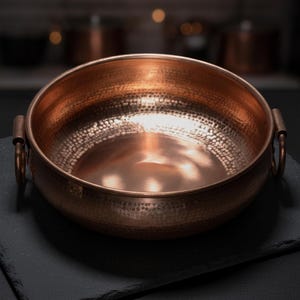 Puede incluir: Un cuenco redondo de cobre martillado con un acabado metálico pulido y dos asas laterales. Las superficies interior y exterior presentan una textura distintiva de hoyuelos que refleja la luz sobre el cálido metal de color marrón rojizo.