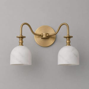 May include: A two-light wall sconce featuring a circular brass backplate and two curved brass arms. Each arm supports a white marble dome-shaped shade with subtle grey veining, connected by a decorative brass spindle detail above each light.