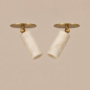 May include: Two ceiling-mounted spotlights featuring cylindrical alabaster shades with natural cream and beige veining. Each light is attached to a circular brass base by a small ball-and-socket joint, allowing for adjustable positioning of the light.