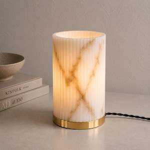 May include: A cylindrical table lamp featuring a translucent, ribbed white body with natural gold-toned veining patterns. The lamp is illuminated from within and sits on a circular, polished gold-colored metal base with a black twisted power cord.