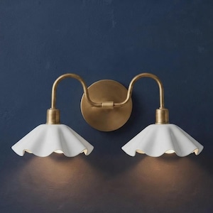 May include: A double wall sconce featuring a circular brass backplate with two curved arms extending outwards. Each arm supports a white, ruffled, bell-shaped shade that directs light downwards, creating a balanced and symmetrical lighting fixture.