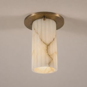 Op de afbeelding: Een cilindrische plafondlamp met een doorschijnende, gebroken witte stenen kap met natuurlijke aders en een verticale geribbelde textuur. De kap is bevestigd aan een ronde, geborsteld messingkleurige metalen voetplaat met een centrale bevestiging.