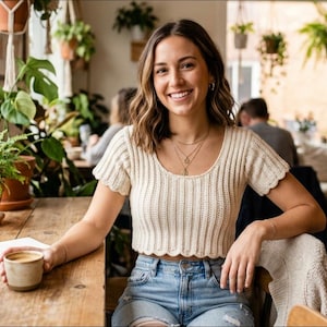 May include: A woman smiles while sitting at a wooden table in a cafe, holding a coffee cup. She wears a cream-colored, short-sleeved, scalloped-hem top and blue denim shorts. Potted plants and hanging plants decorate the cafe.